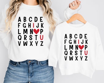 May include: White long-sleeved shirts with black letters spelling out the alphabet. The letter 'I' is replaced with a red heart and the letter 'U' is highlighted in red. The shirts say "I love you" in a unique way.