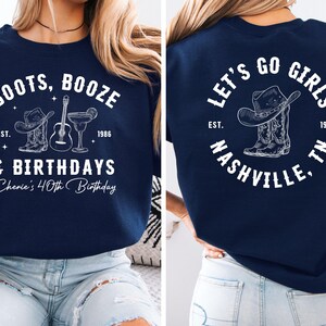 May include: Navy blue sweatshirt with white text and graphics. The front reads "BOOTS, BOOZE & BIRTHDAYS" with a cowboy hat, boots, guitar, and cocktail. The back says "LET'S GO GIRLS NASHVILLE, TN" with a cowboy hat and boots.