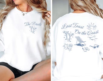 May include: White sweatshirt with blue text and graphics. The front says "The Bride" with two martini glasses. The back says "Last Toast On The Coast" with two martini glasses, a beach chair, seashells, and sunglasses.