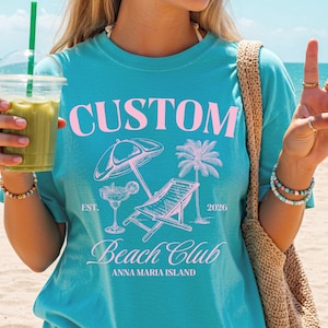 Op de afbeelding: Turquoise t-shirt met een roze opdruk met de tekst "CUSTOM Beach Club ANNA MARIA ISLAND 2026." Het ontwerp bevat een parasol, ligstoel, palmboom en cocktail. De persoon houdt een groen drankje vast.