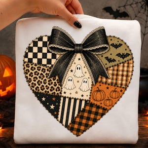 May include: A white sweatshirt featuring a large heart-shaped graphic composed of various patchwork patterns including black and white checkers, leopard print, orange pumpkins, bats, and ghosts, topped with a large black bow at the centre.