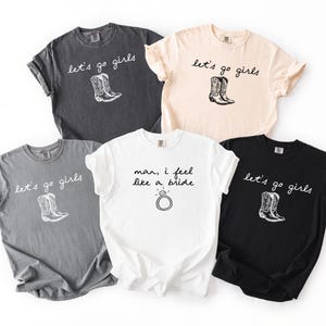 Può includere: Cinque magliette in vari colori: antracite, grigio chiaro, crema, bianco e nero. Ogni maglietta presenta un design grafico. Quattro magliette hanno la scritta "let's go girls" con un'illustrazione di stivale da cowboy. Una maglietta bianca dice "man, I feel like a bride" con un anello.