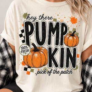 Op de afbeelding: Een crèmekleurig t-shirt met de tekst "hey there PUMPKIN pick of the patch" in het zwart, vergezeld van twee oranje pompoenen, een "100% FRESH" rond embleem en decoratieve groene, oranje en zwarte ruitpatronen en spetters.