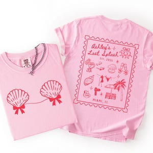 May include: Two pink t-shirts. One features a red seashell design with bows, and a silver chain necklace. The other shirt has a red stamp design with text that reads "Ashley's Last Splash" and "Miami, FL".