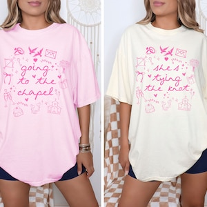 Könnte beinhalten: Zwei übergroße T-Shirts mit Hochzeitsthema. Das rosa Shirt sagt "going to the chapel" mit rosa Hochzeitsmotiven. Das elfenbeinfarbene Shirt sagt "she's tying the knot" mit rosa Hochzeitsmotiven.