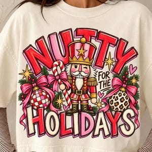 Può includere: Una t-shirt a maniche corte color bianco sporco con una stampa grafica di uno schiaccianoci che tiene un bastoncino di zucchero, circondato da decorazioni natalizie, fiocchi e rami di pino. Il testo "NUTTY FOR THE HOLIDAYS" è in rosso e rosa.