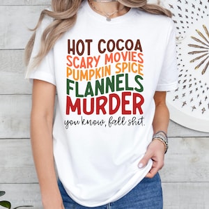 May include: White t-shirt with the words "HOT COCOA, SCARY MOVIES, PUMPKIN SPICE, FLANNELS, MURDER" in various colors. The text "you know, fall shit" is below the list.