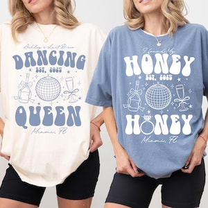 以下が含まれることがあります： レトロなテキストとグラフィックが特徴の、アイボリーとブルーの2枚のTシャツ。アイボリーのシャツには、ディスコボールとシャンパンボトルとともに「Dancing Queen」と書かれています。ブルーのシャツには、指輪とディスコボールとともに「Honey Honey」と書かれています。