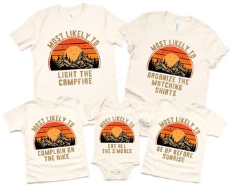 May include: Cream-colored t-shirts and a onesie with a sunset and mountain graphic. The shirts read "Most Likely To" with phrases like "Light the Campfire," "Organize the Matching Shirts," and "Eat All the S'mores."