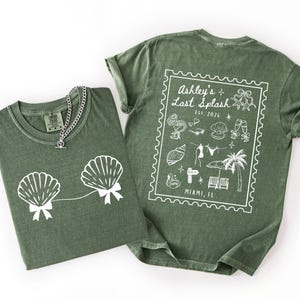 May include: Two olive green t-shirts with white graphics. One shirt features seashell designs with bows, and a silver chain necklace. The other shirt has a stamp-like design with text that reads "Ashley's Last Splash" and "Miami, FL".