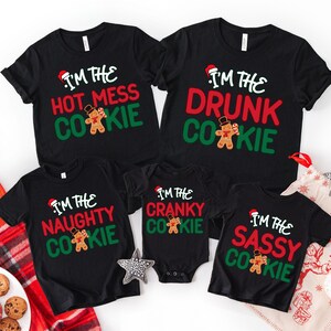 May include: A collection of black Christmas-themed t-shirts and a onesie. Each shirt features the phrase "I'm the" followed by a different adjective and "Cookie", along with a gingerbread man graphic and a Santa hat. The phrases include "Hot Mess", "Drunk", "Naughty", "Cranky", and "Sassy".
