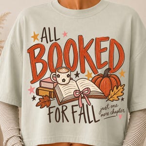 May include: A light sage green t-shirt featuring a graphic print with the text ALL BOOKED FOR FALL and just one more chapter. The design includes an open book with a pink ribbon, a stack of books, a coffee mug, a pumpkin, leaves, and stars.