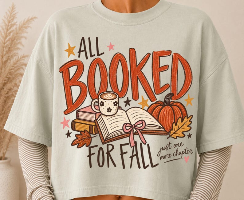 May include: A light sage green t-shirt featuring a graphic print with the text ALL BOOKED FOR FALL and just one more chapter. The design includes an open book with a pink ribbon, a stack of books, a coffee mug, a pumpkin, leaves, and stars.