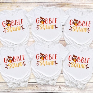 May include: Six white t-shirts with the text "GOBBLE SQUAD" in red and yellow, featuring a cartoon turkey. The shirts are arranged on a light wood-grain surface. The shirts are tied in a knot at the bottom.
