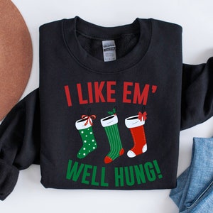 Op de afbeelding: Zwart sweatshirt met de tekst "I LIKE EM' WELL HUNG!" in rood en groen. Het ontwerp toont drie kerstsokken in groen, rood en wit met decoratieve strikken. Een feestelijk kledingstuk.