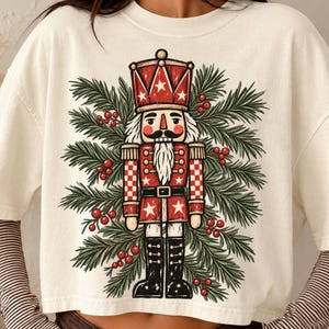 Peut inclure: Un t-shirt à manches courtes blanc cassé orné d'un motif de soldat casse-noisette en tenue rouge et blanche, entouré de branches de pin vertes et de baies rouges. Le t-shirt est superposé sur un haut à manches longues à rayures marron et blanches.