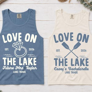 May include: A blue tank top with white text reading LOVE ON THE LAKE, EST. 2026, Future Mrs. Taylor, LAKE TAHOE, and a diamond ring graphic, alongside a cream tank top with blue text reading LOVE ON THE LAKE, EST. 2026, Casey's Bachelorette, LAKE TAHOE.