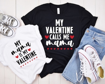 Pode incluir: Duas camisetas com texto temático do Dia dos Namorados. A camiseta branca diz "My mama is my Valentine" com corações vermelhos. A camiseta preta diz "My Valentine calls me mama" com corações vermelhos.