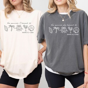 May include: Two oversized t-shirts, one cream and one charcoal gray, each with a white graphic. The graphic includes the text "the summer I turned 30" and "MIAMI, FL 2026" along with seashell, palm tree, and cocktail illustrations.