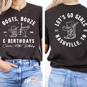 May include: Dark grey t-shirt with white text and illustrations. The front says "BOOTS, BOOZE & BIRTHDAYS" with a cowboy hat, boots, guitar, and cocktail graphic. The back says "LET'S GO GIRLS NASHVILLE, TN" with a hat and boots graphic.