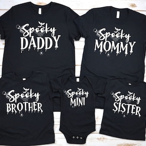Puede incluir: Camisetas negras con texto blanco que dice "Spooky Daddy", "Spooky Mommy", "Spooky Brother", "Spooky Mini" y "Spooky Sister". Cada camiseta tiene un diseño de Halloween con murciélagos, telarañas y detalles goteantes.