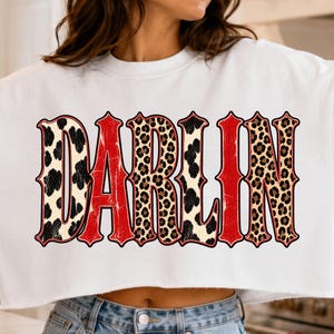 Peut inclure: Un sweat-shirt court blanc avec le mot DARLIN imprimé en grandes lettres stylisées. Les lettres alternent entre une texture rouge vieillie et un motif léopard marron et noir, créant un design graphique audacieux sur le devant.