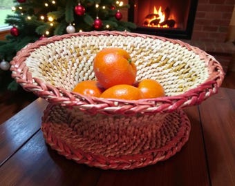 Puede incluir: Una cesta tejida con base de pedestal, llena de varias naranjas. La cesta tiene un tejido natural de color claro con un borde rojo. El fondo incluye un árbol de Navidad y una chimenea.