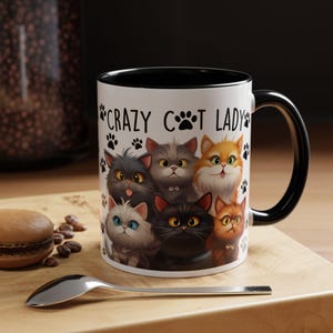 May include: White ceramic mug with a black rim and handle. The mug features a design of six cartoon cats with the text "Crazy Cat Lady" and paw prints.