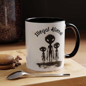 May include: A white ceramic mug with a black interior and handle. The mug features the text "Illegal Aliens!" above a graphic of three alien figures. A spoon and a macaron are in the foreground.