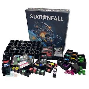 Op de afbeelding: Een zwart-witte gamebox met de titel "Stationfall" op de voorkant. De doos is omringd door verschillende zwarte plastic containers gevuld met spelstukken, dobbelstenen en kaarten. De containers zijn zo georganiseerd dat ze op een game-organizer wijzen.
