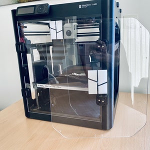 K&ouml;nnte beinhalten: Ein schwarzer 3D-Drucker mit einer transparenten Plastikt&uuml;r und einer transparenten Plastikseitenteil. Der Drucker tr&auml;gt den Text "Bambu Lab P1S" auf der Vorderseite.