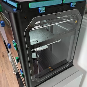 K&ouml;nnte beinhalten: Ein schwarzer 3D-Drucker mit einer durchsichtigen Plastikt&uuml;r und einem silbernen Metallrahmen. Der Drucker hat einen blauen und gr&uuml;nen Streifen oben und das Wort "ARCO" auf der Vorderseite.