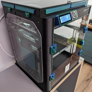 K&ouml;nnte beinhalten: Ein schwarzer 3D-Drucker mit einem Glaseinschluss und einem digitalen Display. Der Drucker steht auf einem Holztisch mit einer schwarzen Matte.