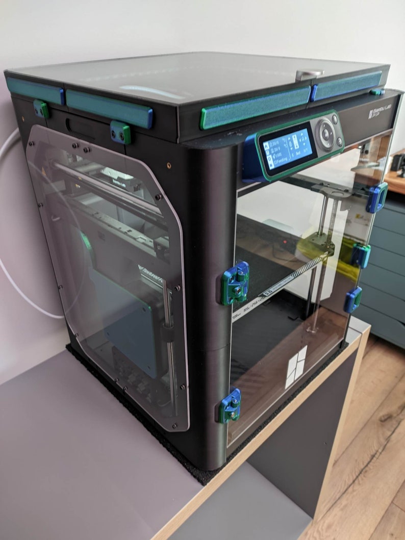 K&ouml;nnte beinhalten: Ein schwarzer 3D-Drucker mit einem Glaseinschluss und einem digitalen Display. Der Drucker steht auf einem Holztisch mit einer schwarzen Matte.