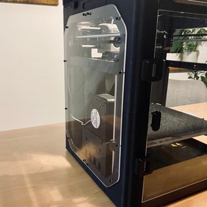 K&ouml;nnte beinhalten: Schwarzer 3D-Drucker mit einer durchsichtigen Plastikt&uuml;r und einem sichtbaren L&uuml;fter im Inneren. Der Drucker steht auf einer Holzoberfl&auml;che.