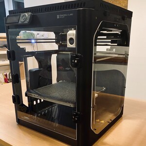K&ouml;nnte beinhalten: Ein schwarzer 3D-Drucker mit einem Glaseinschluss. Der Drucker hat ein Touchscreen-Display und eine Kamera oben. Der Drucker steht auf einem Holztisch.