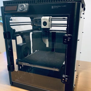 K&ouml;nnte beinhalten: Ein schwarzer 3D-Drucker mit einem transparenten Kunststoffgeh&auml;use. Der Drucker hat eine schwarze Basis und eine wei&szlig;e Kamera oben. Der Drucker ist mit "Bambu Lab P1S" beschriftet.