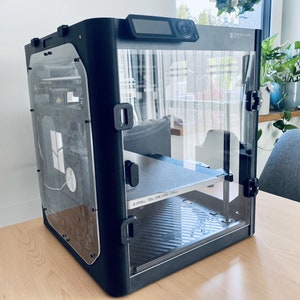 K&ouml;nnte beinhalten: Ein schwarzer 3D-Drucker mit einem transparenten Kunststoffgeh&auml;use. Der Drucker hat einen kleinen LCD-Bildschirm an der Vorderseite und eine Bauplattform im Inneren. Der Drucker steht auf einem Holztisch.
