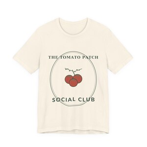 Può includere: Una t-shirt a maniche corte color crema con una grafica centrale di tre pomodori rossi su un rametto racchiusi in un sottile contorno ovale verde scuro. Il testo THE TOMATO PATCH è stampato sopra e SOCIAL CLUB sotto.