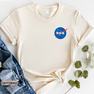 Op de afbeelding: Een cr&egrave;mekleurig T-shirt met een blauw en rood NASA-ge&iuml;nspireerd logo met de tekst "MOM".