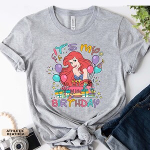Puede incluir: Una camiseta gris con un gr&aacute;fico colorido de Ariel de La Sirenita. El gr&aacute;fico incluye un pastel de cumplea&ntilde;os, regalos y globos. El texto "Es mi cumplea&ntilde;os" est&aacute; escrito en la camiseta.
