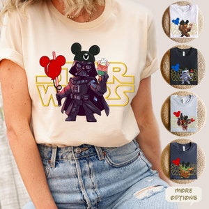 May include: A white t-shirt with a yellow "Star Wars" graphic featuring Darth Vader holding a cupcake and a red balloon. Darth Vader is wearing a Mickey Mouse hat. The shirt is being modeled by a person wearing blue jeans.