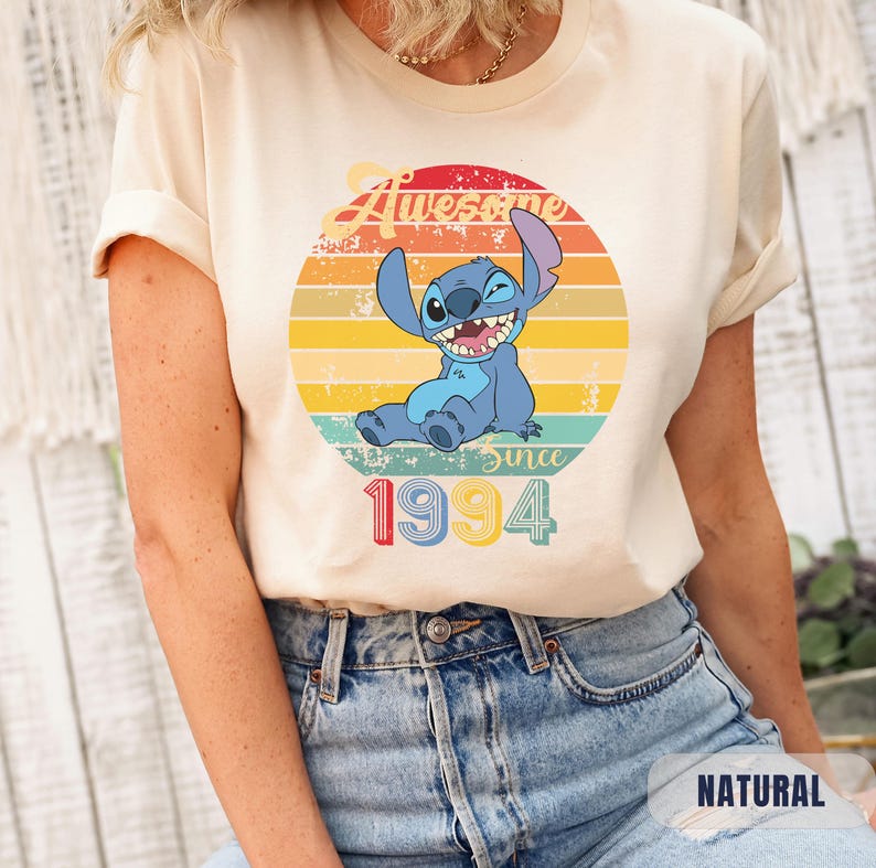 May include: A light beige t-shirt with a vintage-style graphic of a blue cartoon character with a winking eye. The graphic includes the text "Awesome since 1994" and a rainbow-colored sunset.