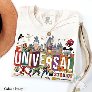 May include: Ivory-colored t-shirt featuring a colorful graphic of the Universal Studios logo with various cartoon characters. The shirt includes characters from Marvel, Harry Potter, and other popular franchises. A gold necklace and a brown hat are also visible.