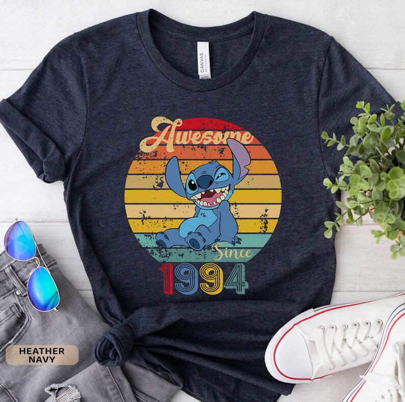 May include: A navy blue t-shirt with a vintage-style graphic of Stitch from Lilo & Stitch. The graphic features Stitch with a mischievous grin, sitting in a sunset with the text "Awesome Since 1994".