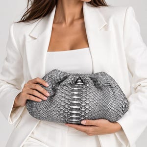 Pode incluir: Uma bolsa clutch com padrão de pele de cobra em cinzento e branco, com um fecho franzido na parte superior. A bolsa estilo bolsa apresenta uma superfície texturizada semelhante a escamas e uma silhueta arredondada, segurada horizontalmente.