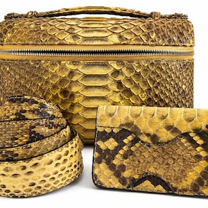 May include: A yellow and brown snakeskin patterned vanity case with a top handle and zipper closure, a coiled snakeskin belt, and a rectangular snakeskin wallet. The set features a consistent scale texture in shades of mustard yellow and dark brown.