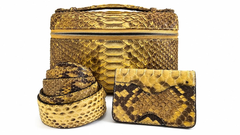 May include: A yellow and brown snakeskin patterned vanity case with a top handle and zipper closure, a coiled snakeskin belt, and a rectangular snakeskin wallet. The set features a consistent scale texture in shades of mustard yellow and dark brown.