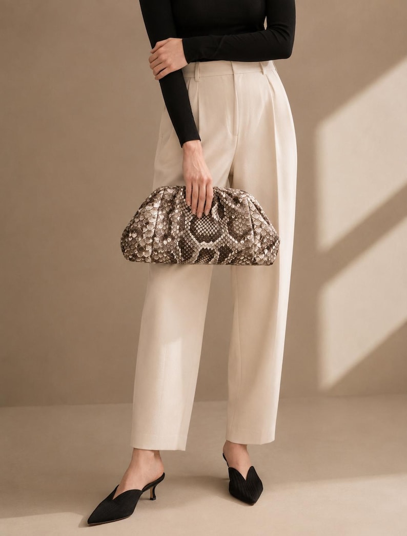 May include: A long-sleeved black top, cream-colored high-waisted trousers with pleats, and black pointed-toe mules. A brown and cream snakeskin-patterned clutch bag with a gathered, rounded frame is held in front of the trousers.