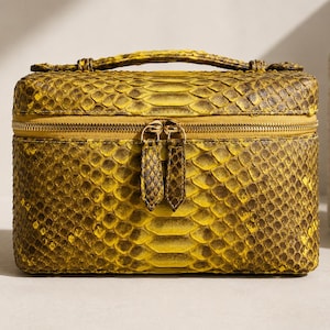 May include: A rectangular vanity case featuring a vibrant yellow and dark brown snakeskin pattern. The structured bag includes a top handle, a gold-toned zipper closure with two matching snakeskin-textured pull tabs, and a boxy silhouette.